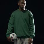 Studio portrait of an African American man in a green sweatshirt holding a soccer ball.