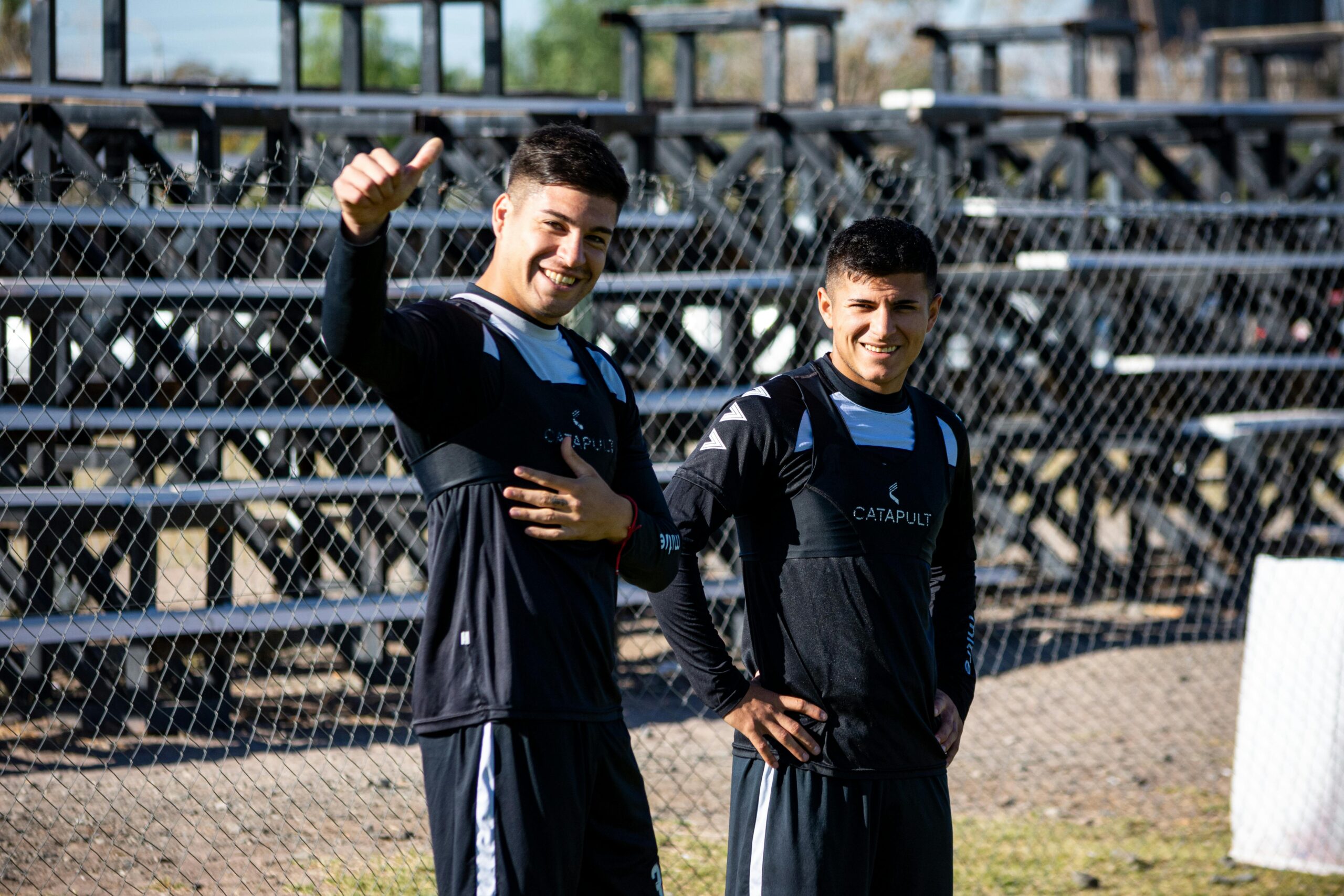 Two young soccer players in training gear smile cheerfully during practice on a sunny day.